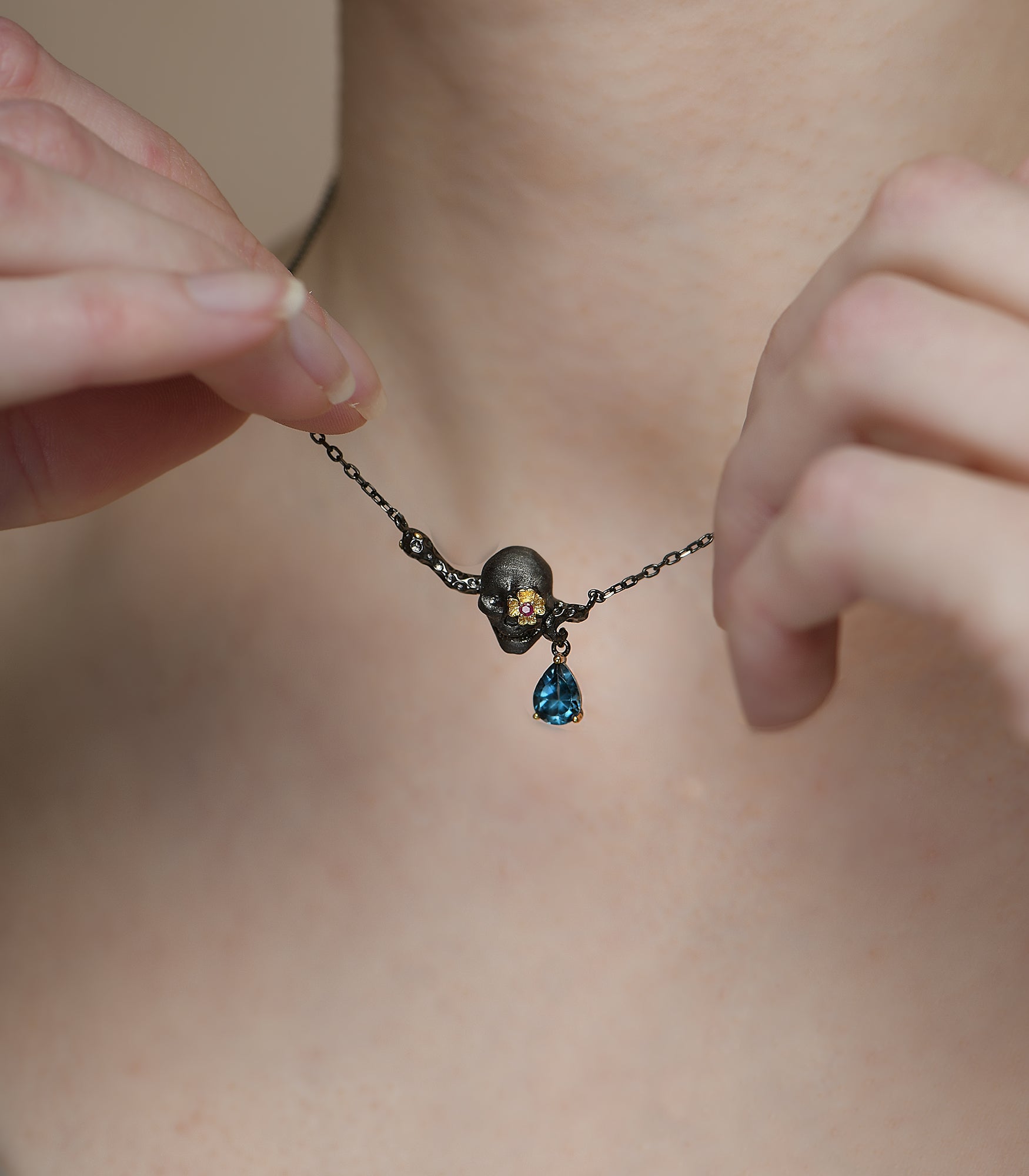 An oxidised sterling silver necklace featuring a skull pendant with a gold vermeil flower, pink ruby and blue topaz gemstone.