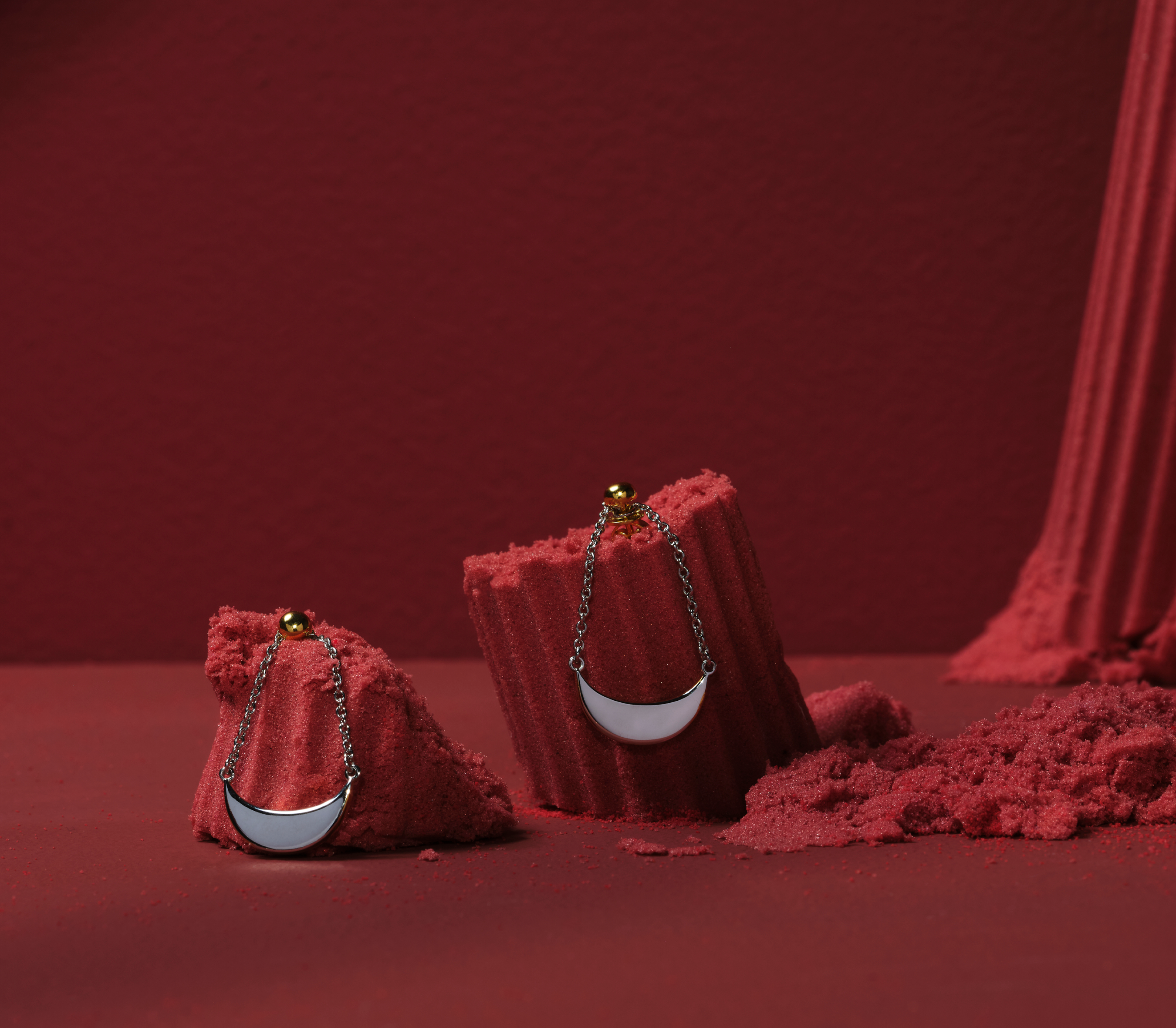 A pair of earrings are hung in front of a red background. The earrings have 2 dainty chains holding a half crescent moon.