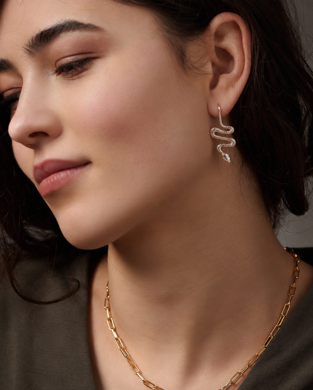 A close-up of a woman wearing a delicate silver snake-shaped drop earring and a gold chain necklace, shown in profile against a soft gray background.