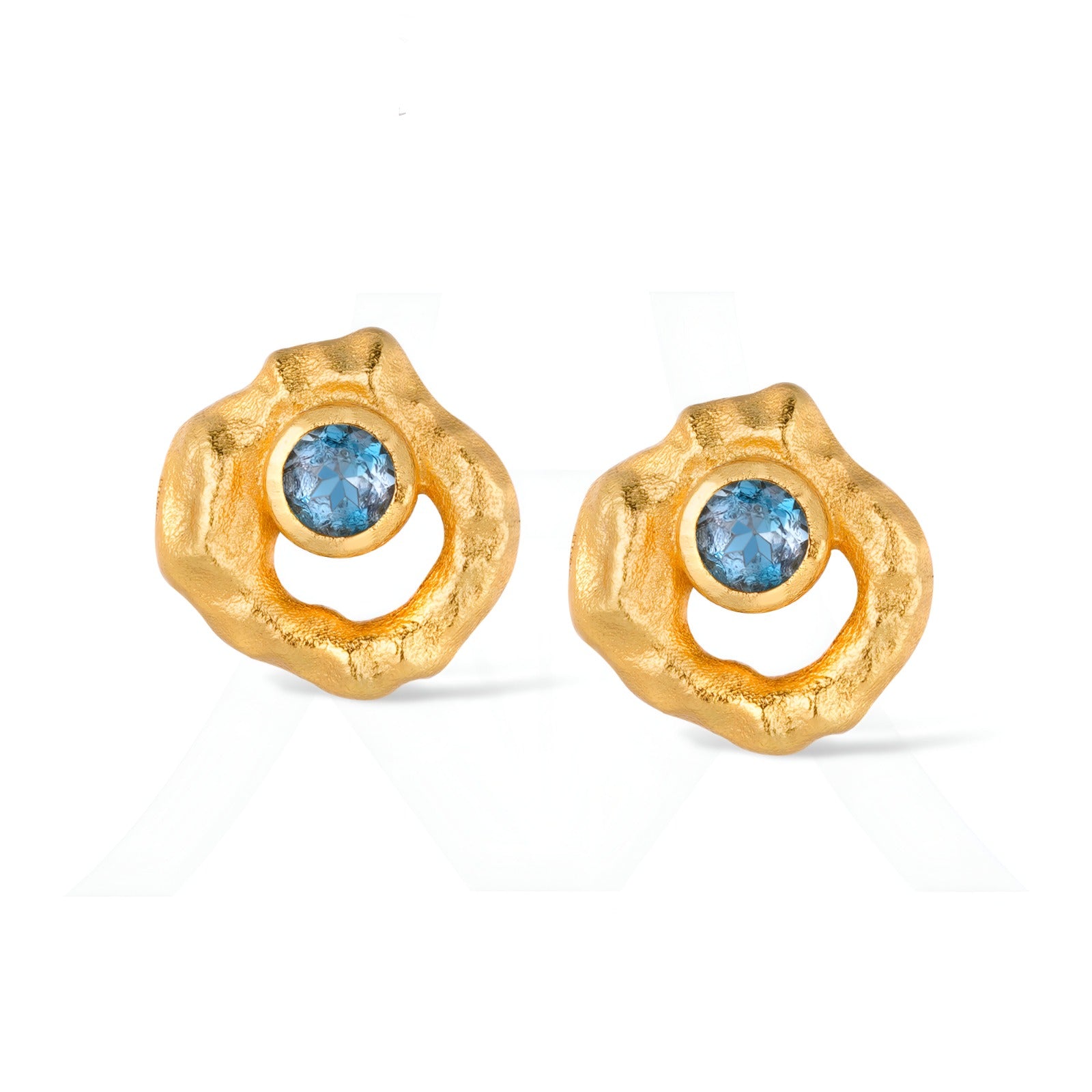 Pair of textured hammered gold stud earrings with a central bezel-set round blue topaz or sapphire.

Close-up of artisan organic-shaped gold earrings with blue gemstones