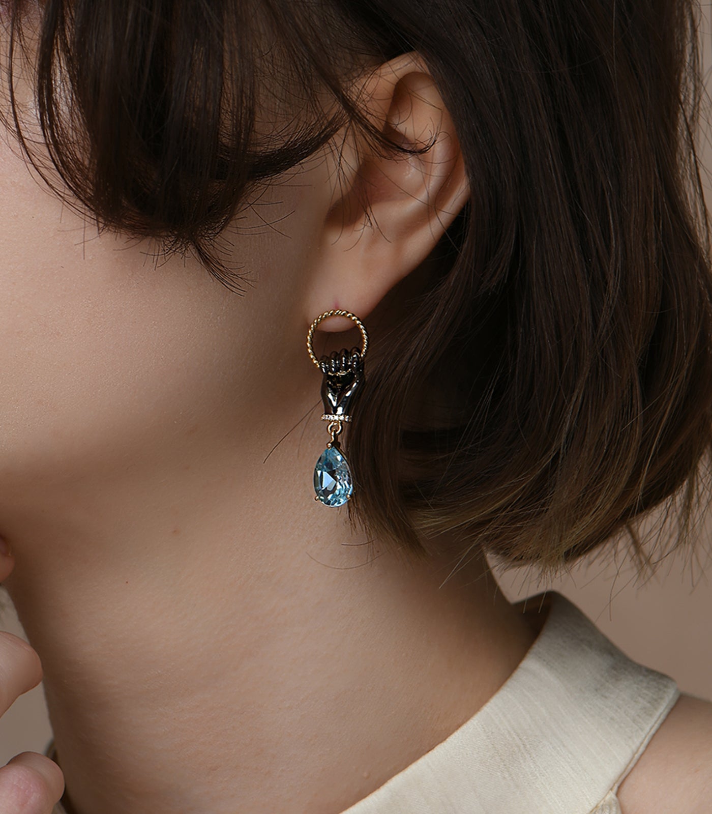 A pair of earrings with a small hoop stud and a fist held on to the hoop. A blue topaz stone dangles from the hand.