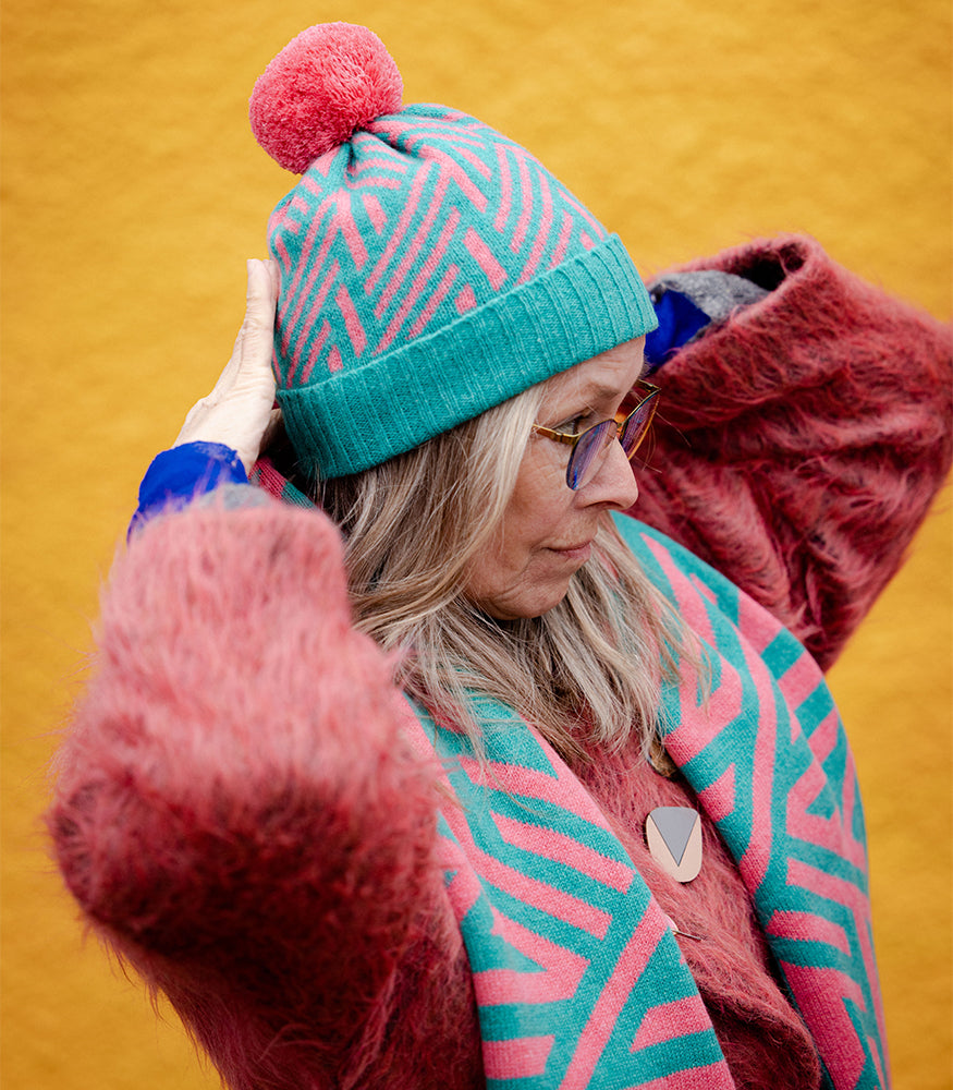 A lady wearing a pink and blue bobble hat with a matching scarf. The hat has a geometric crosswise pattern.