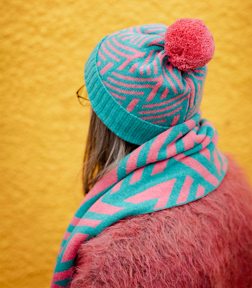 A lady wearing a pink and blue bobble hat with a matching scarf. Both the hat and scarf have a matching crosswise pattern.