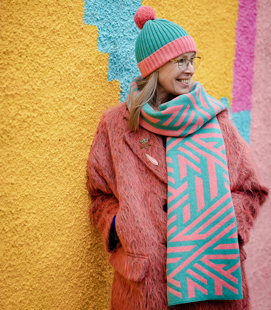 A lady wearing a pink and blue bobble hat with a matching scarf. The scarf has a geometric crosswise pattern.