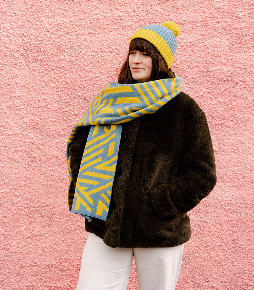 A lady wearing a cosy yellow and blue scarf with a geometric crosswise pattern. The scarf is paired with a matching hat.