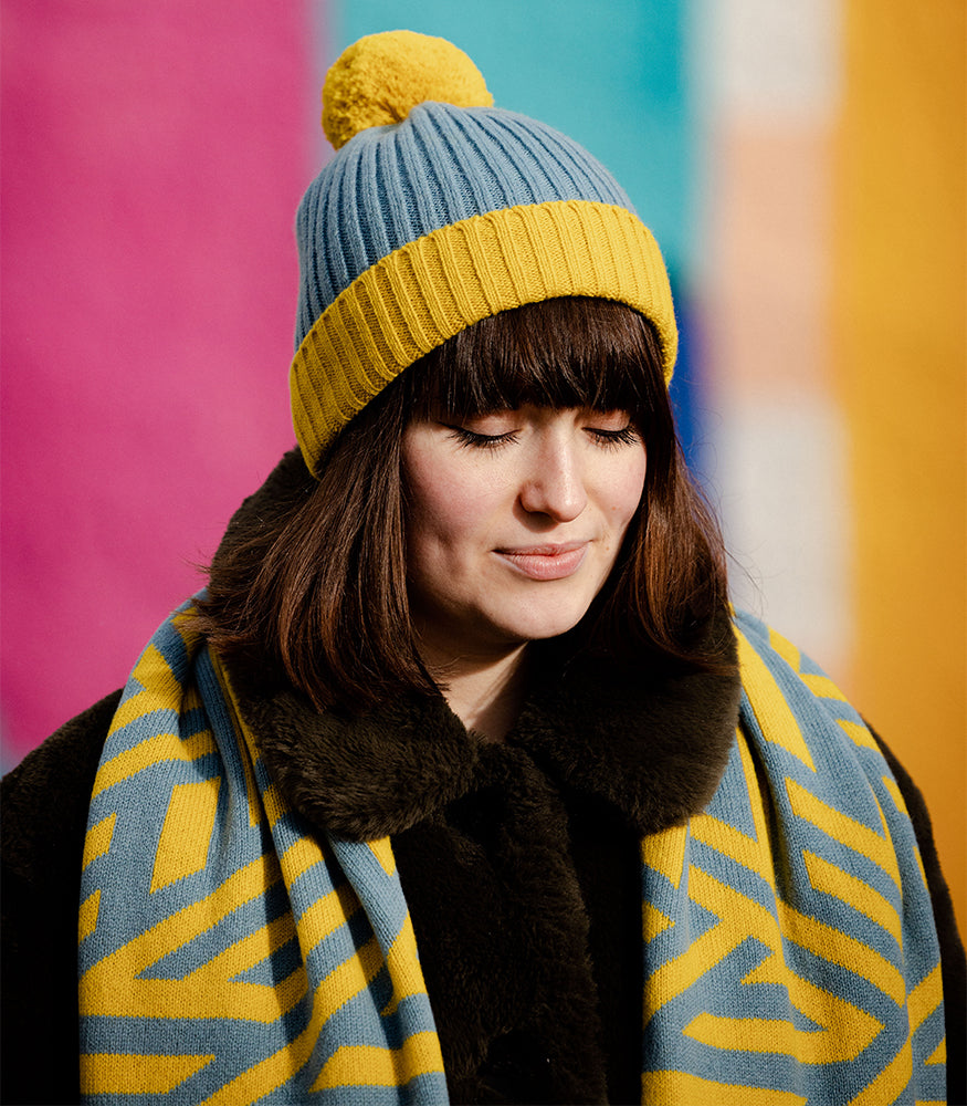 A woman wearing a ribbed beanie bobble hat. The beanie is yellow and blue.