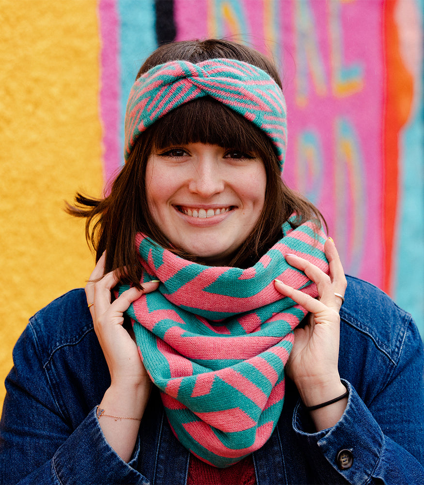 A woman wearing a pink and blue snood with a geometric crosswise pattern. The woman also wears a matching twist headband.