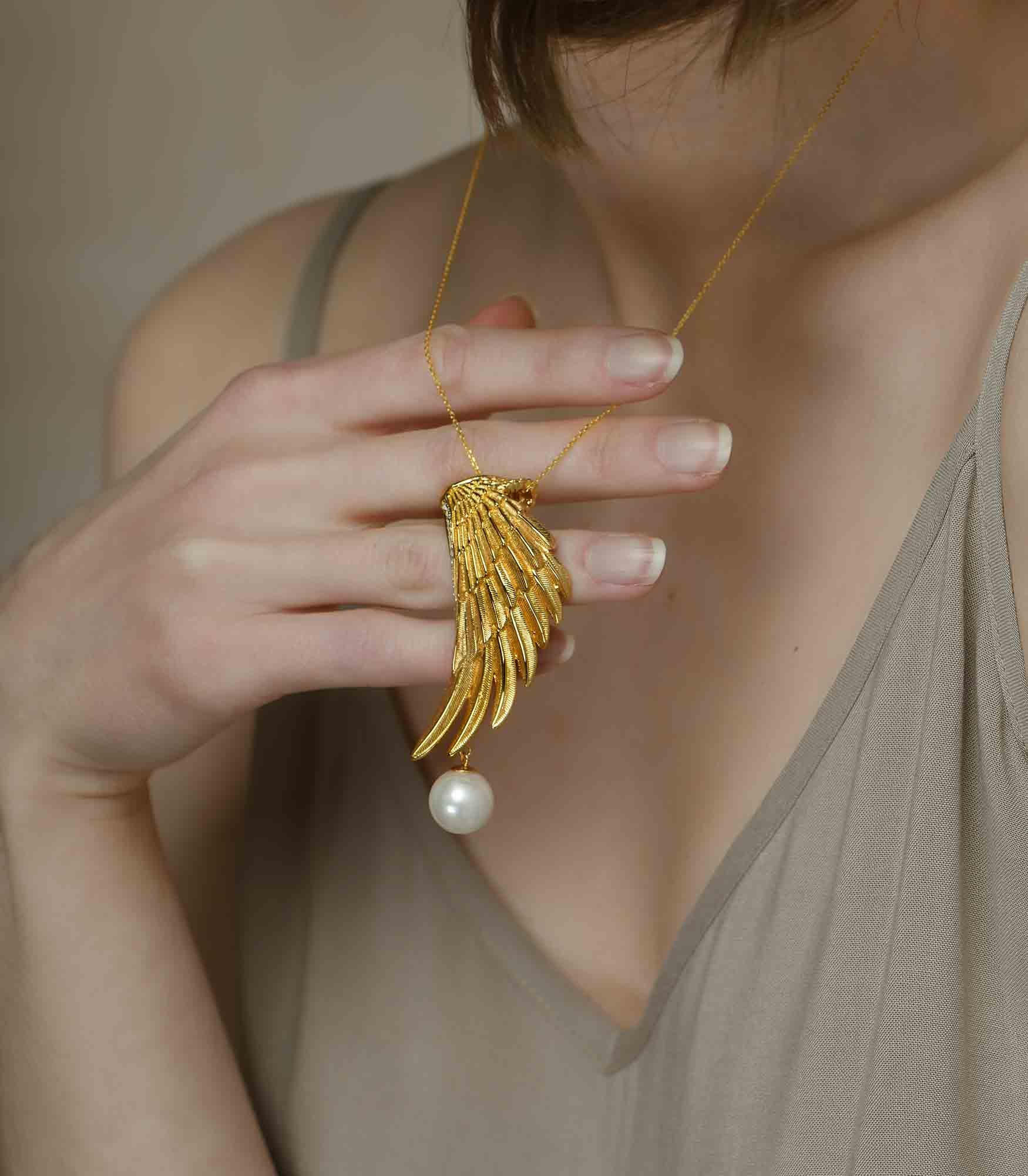 A lady holding a gold vermeil necklace which has an eagle wing pendant with a white pearl hanging from it.