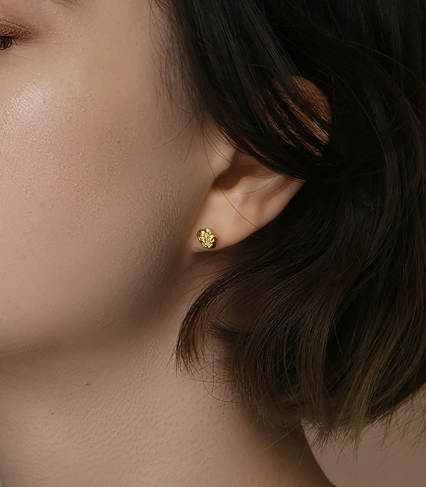 A model wears a pair of gold plated stud earrings. The earrings are a pair of small textured nuggets.