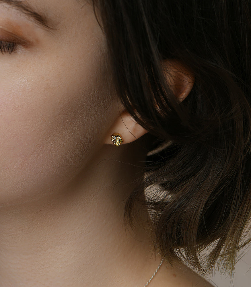 A model wears a pair of gold plated earrings. The earrings are a pair of small, textured nugget studs.