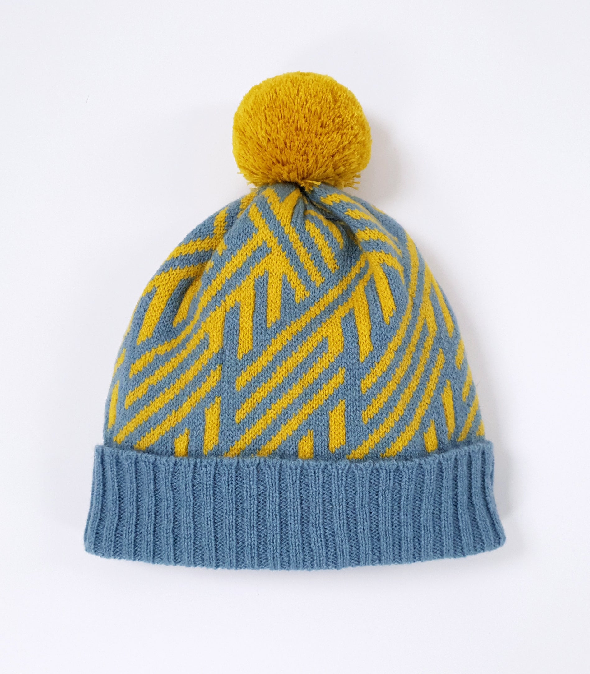 A knitted pom pom beanie hat with a ribbed blue hem. The main body of the hat has a yellow and blue crosswise pattern.