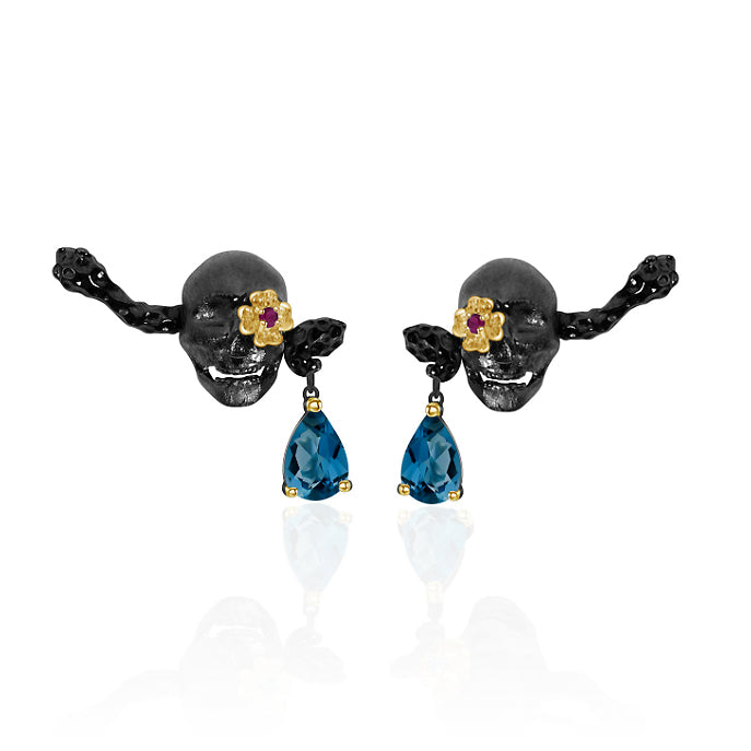 A pair of oxidised black skull earrings. The earrings feature two blue topaz and pink ruby stones, injecting some colour into the design.